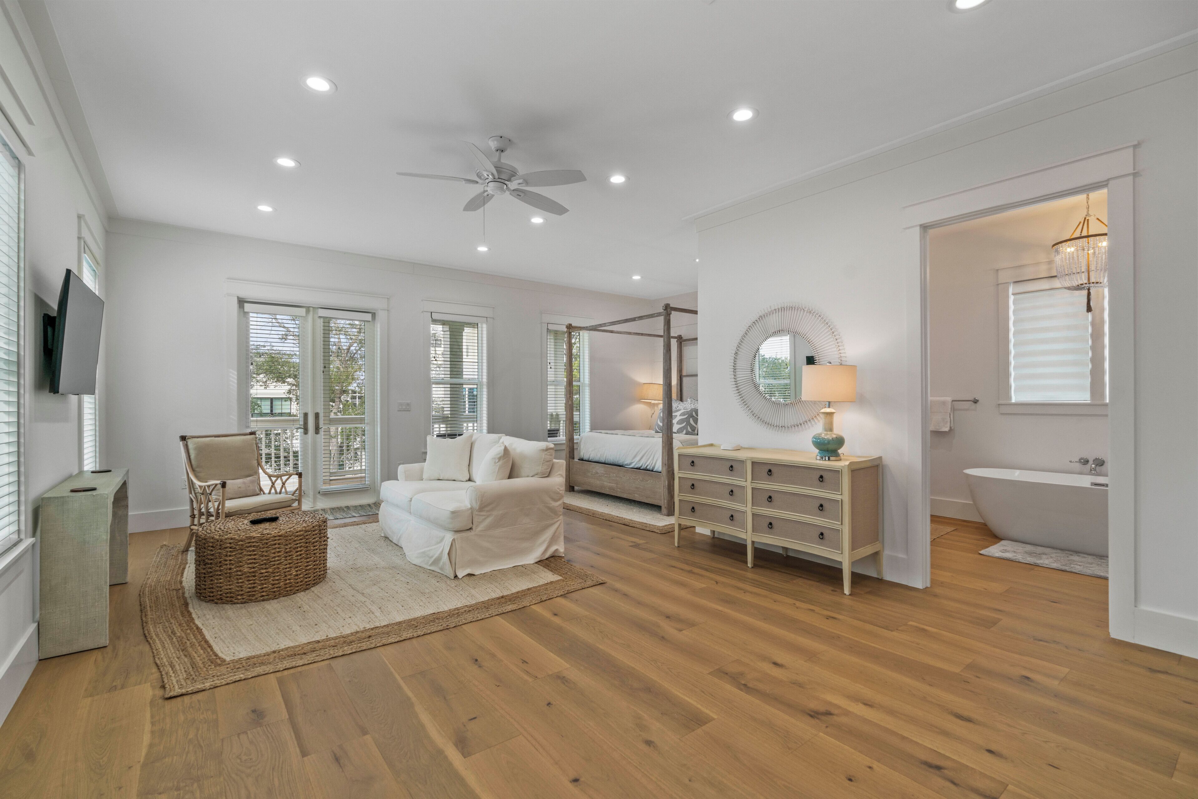 A spacious bedroom with a large bed, a vanity with a mirror, a ceiling fan, and a view of the outdoors through the windows.