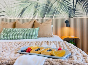 A neatly made bed with a fluffy beige blanket, a plate of fruit and pastries, and a wall with vertical blinds and a lamp. - NEW! 3 Pools | Tiki Bar | 2nd Floor | Sleeps 10 | Renovated! (Panama City Beach)
