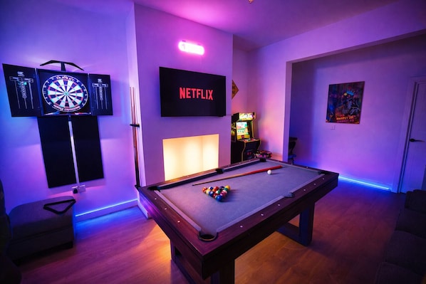 A pool table with billiard balls, a dartboard, a TV screen with 'NETFLIX' logo, and a poster on the wall.