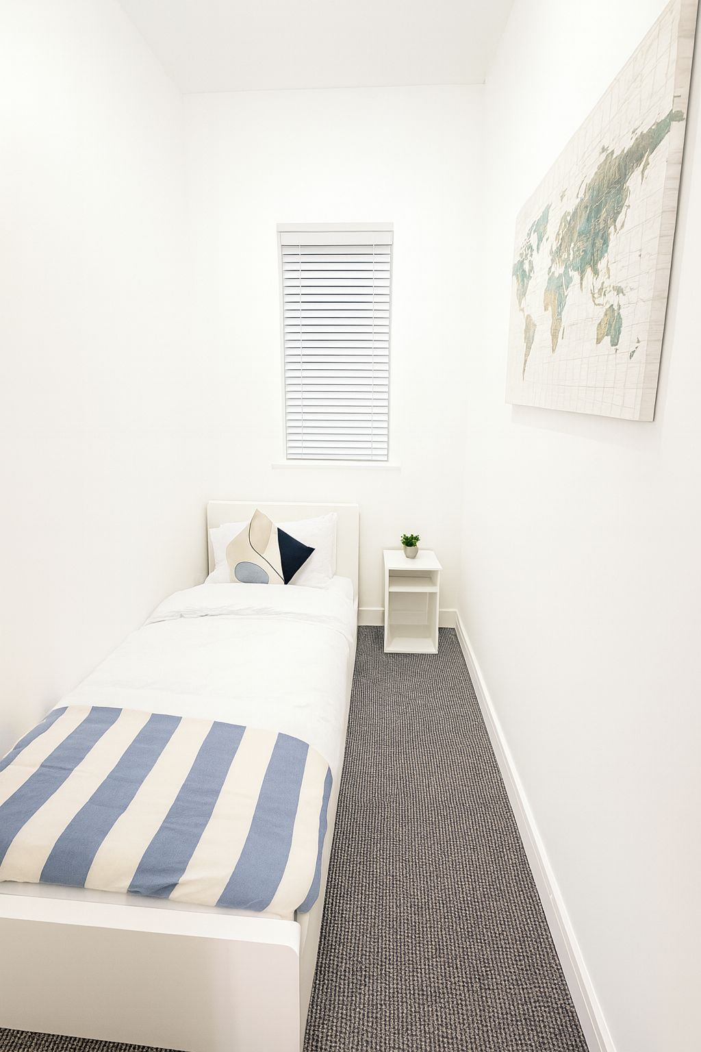 A hotel room with a bed, a small bedside table with a plant, a window with blinds, and a world map on the wall.