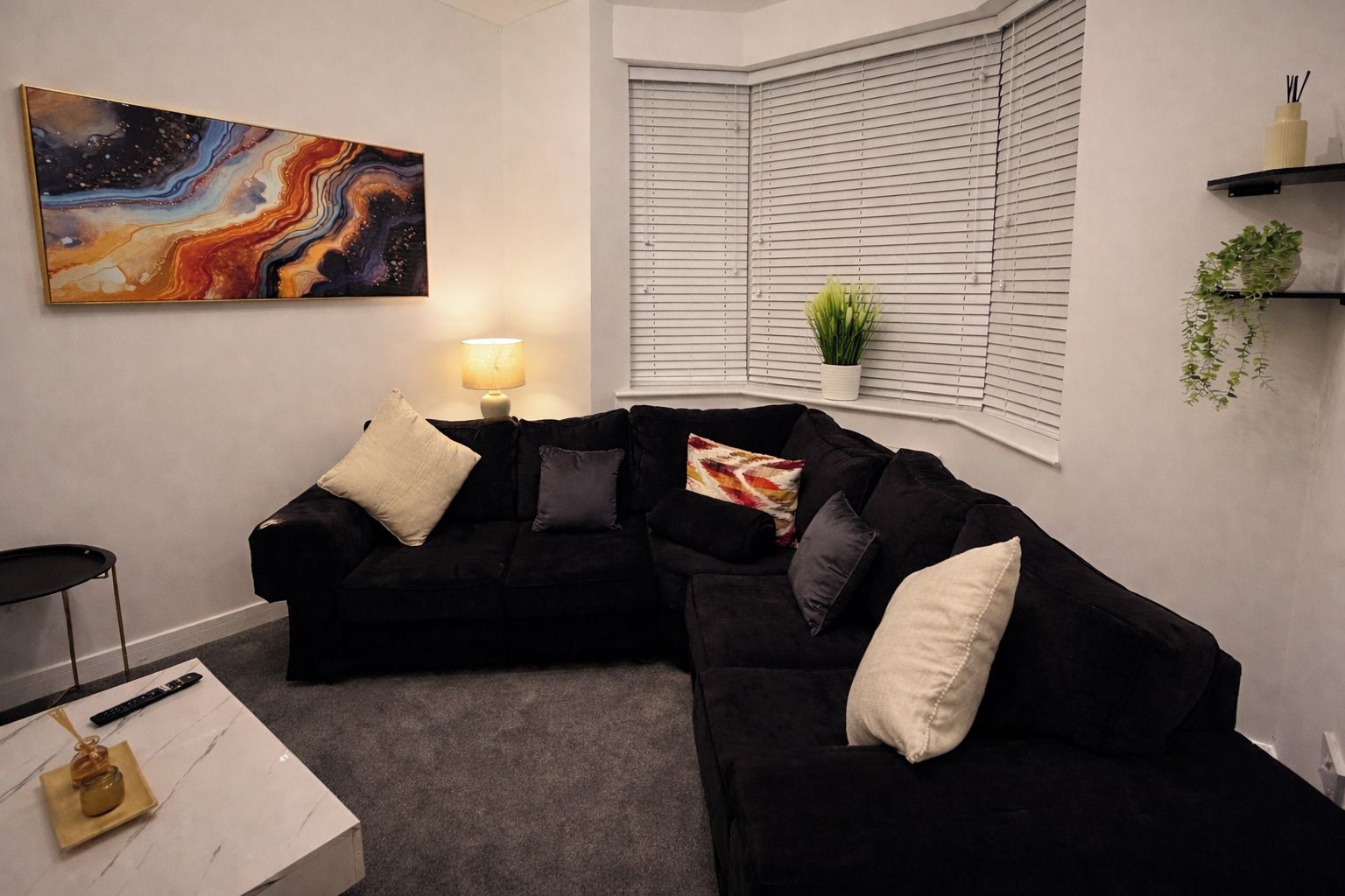 Un salon moderne avec un canapé d’angle noir, une table basse blanche, un tableau au mur, une fenêtre avec des stores et une petite plante posée sur une étagère.