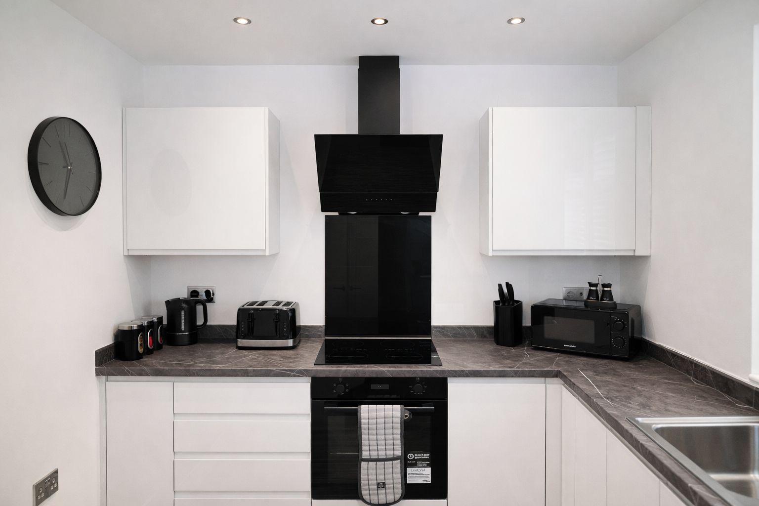 A modern kitchen with white cabinets, a black range hood, a microwave, a toaster, and a clock on the wall.