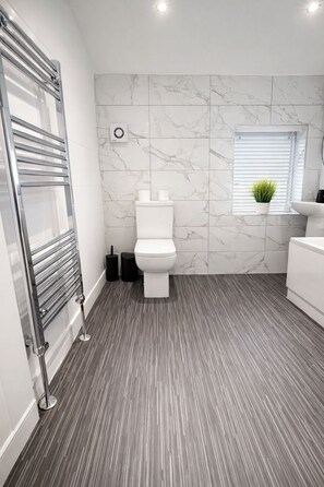 Un baño con un inodoro blanco, un perchero, una planta en maceta y una ventana con persianas.