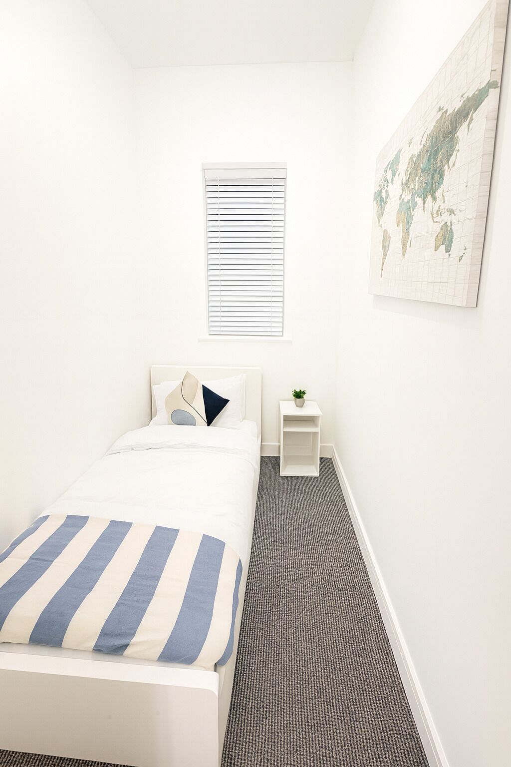 A bedroom with a bed, a small bedside table with a plant, a window with blinds, and a world map on the wall.
