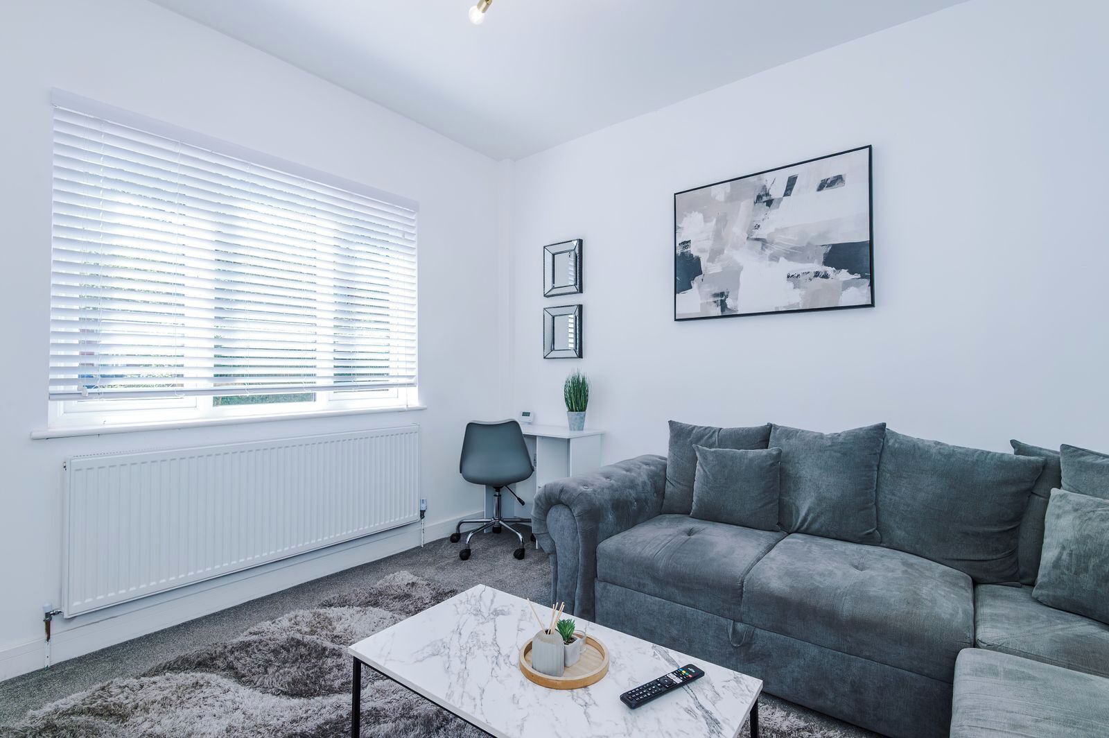 Un salon moderne avec un canapé gris, une table basse blanche et une grande fenêtre équipée de stores.