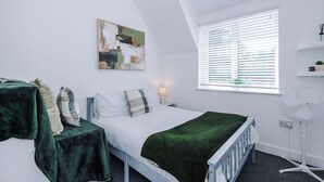 A bedroom with a bed, bedside tables, a window with blinds, a wall-mounted artwork, and a standing fan.