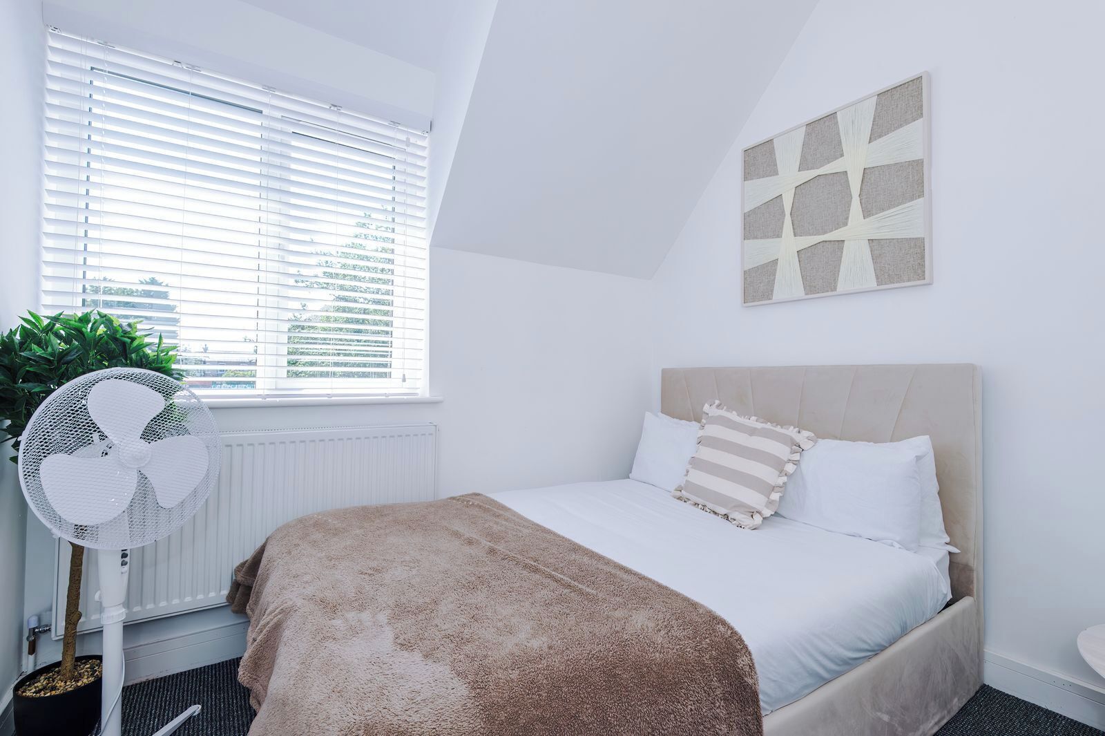 Quarto com uma cama, radiador, janela com persianas, ventilador e uma planta.
