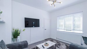 A modern living room with a flat-screen TV, grey sofas, a coffee table, and a large window with blinds.