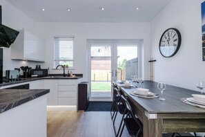 A modern kitchen with a dining area, a large wall clock, and a view through a glass door. - Modern Home for 9 with Garden (Greater Manchester)