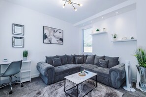 Een moderne woonkamer met een grijze modulaire bank, een witte salontafel, een bureau met stoel, en een groot abstract schilderij.
