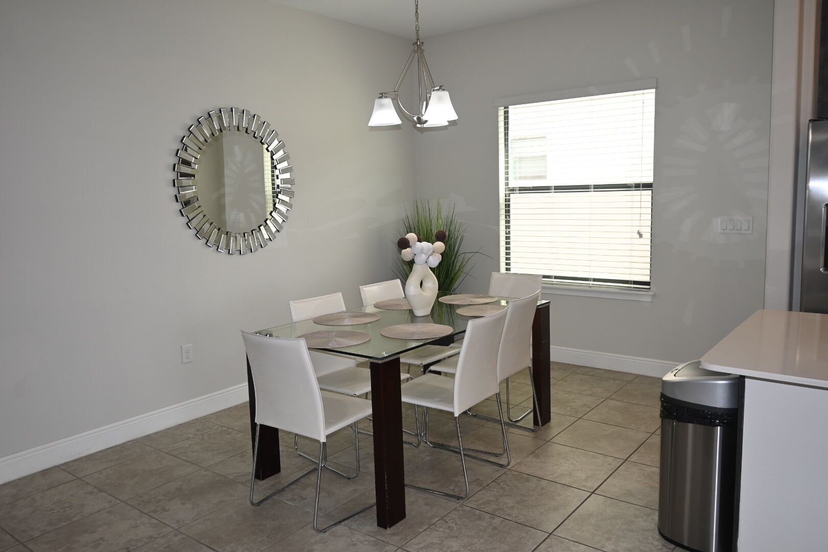A dining area with a glass table, white chairs, a modern mirror, and a window with blinds.