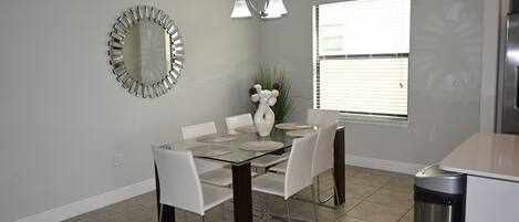 A dining area with a glass table, white chairs, a modern mirror, and a window with blinds.