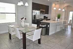 A modern kitchen with a central island, stainless steel appliances, and a dining area with a glass table and white chairs. - Lakeview 4BR/4.5BA Retreat w/ Private Pool Near Legoland & Disney (Haines City)