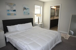 A bedroom with a bed, nightstand, artwork, window, and a bathroom visible through an open door. - Lakeview 4BR/4.5BA Retreat w/ Private Pool Near Legoland & Disney (Haines City)
