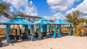 Ein Poolbereich mit mehreren blauen Sonnensegeln, Liegestühlen und einer gefliesten Terrasse.