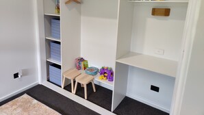 Quarto com roupeiro embutido, um banquinho de madeira e estantes com livros e brinquedos.