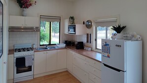 Uma cozinha com armários brancos, fogão, geladeira, janela com vista para o verde e um relógio na parede.