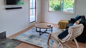 Sala de estar com televisão de tela plana, ventilador de teto, sofá, poltrona de balanço, mesa de centro e uma grande janela com vista para o verde.