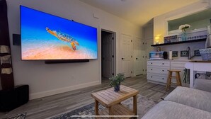 Ein Wohnzimmer mit Flachbildfernseher, einem Holztisch, einer grauen Couch, einer Küchenzeile mit Mikrowelle und einem Flur.