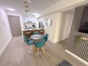 A modern kitchen with white cabinets, a dining area with a round table and teal chairs, and a striped wall with a TV. - Departamento en Miraflores con Piscina (1003). (Lima)