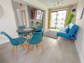A modern living room with a blue sofa, a dining table with four chairs, a large window with a view of buildings, and a potted plant. - Departamento en Miraflores con Piscina (1003). (Lima)