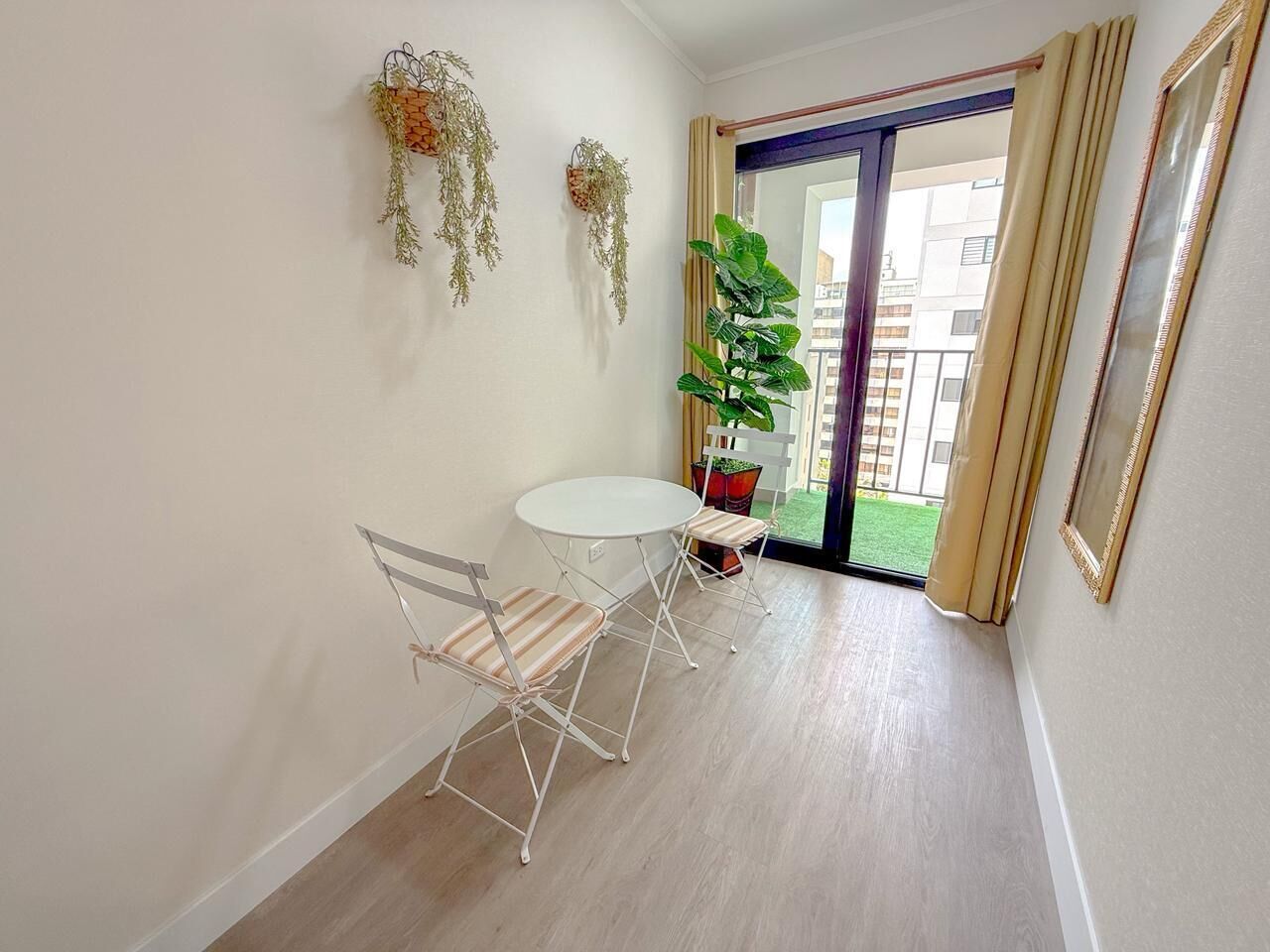 Una habitación con una mesa y sillas blancas, una planta en maceta y un ventanal corredero que da a un balcón.