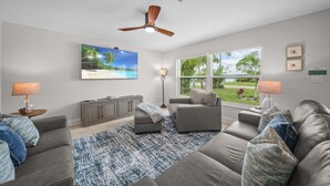A living room with a ceiling fan, a large TV, a window with a view of greenery, and a gray sofa. - Naples Paradise - Heated Saltwater Pool/Spa, Walk/Bike to the Beach! (Naples)
