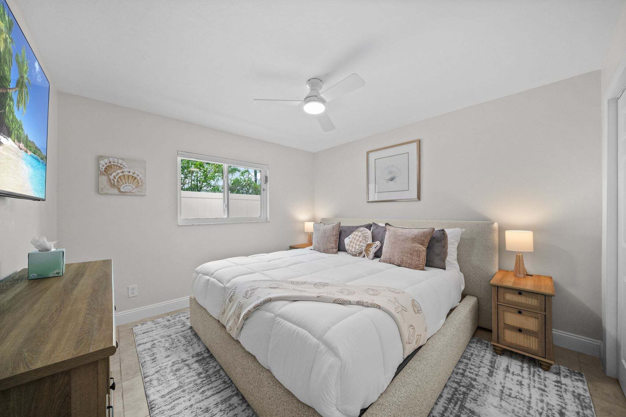 Une chambre à coucher comprenant un lit, une table de chevet, un bureau, un ventilateur de plafond, une fenêtre et des œuvres d’art mural.