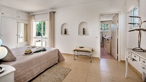 Een slaapkamer met een bed, nachtkastje en een bureau met lades. Er is een raam met gordijnen, een deur naar een badkamer en een klein bankje met een boek.