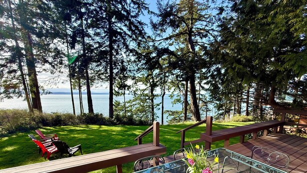 A deck with glass-top tables and metal chairs, overlooking a grassy area with trees and a body of water. - NEW LISTING-Mutiny Bay Ebb Tide Cabin AMAZING SUNSETS, EAGLES GALORE SANDY BEACH (Freeland)