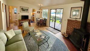 A living room with a sofa, a glass coffee table, a fireplace, and a dining area with chairs and a table. - NEW LISTING-Mutiny Bay Ebb Tide Cabin AMAZING SUNSETS, EAGLES GALORE SANDY BEACH (Freeland)