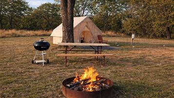 Un camping con tienda de campaña, fogón, parrilla y mesa de picnic.