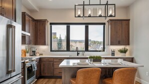 A modern kitchen with stainless steel appliances, wooden cabinets, a large island with two chairs, and a view through a large window. - Downtown WP Loft 57 I Rooftop Hot Tub & Lounge I Great Views I Bunk Room I Discounted Attractions (Winter Park)