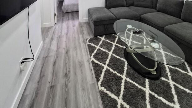A modern living room with a grey sectional sofa, a glass coffee table, and a TV mounted on the wall. - Quaint 2-bedroom house in Chestermere perfect for relaxing stays (Chestermere)