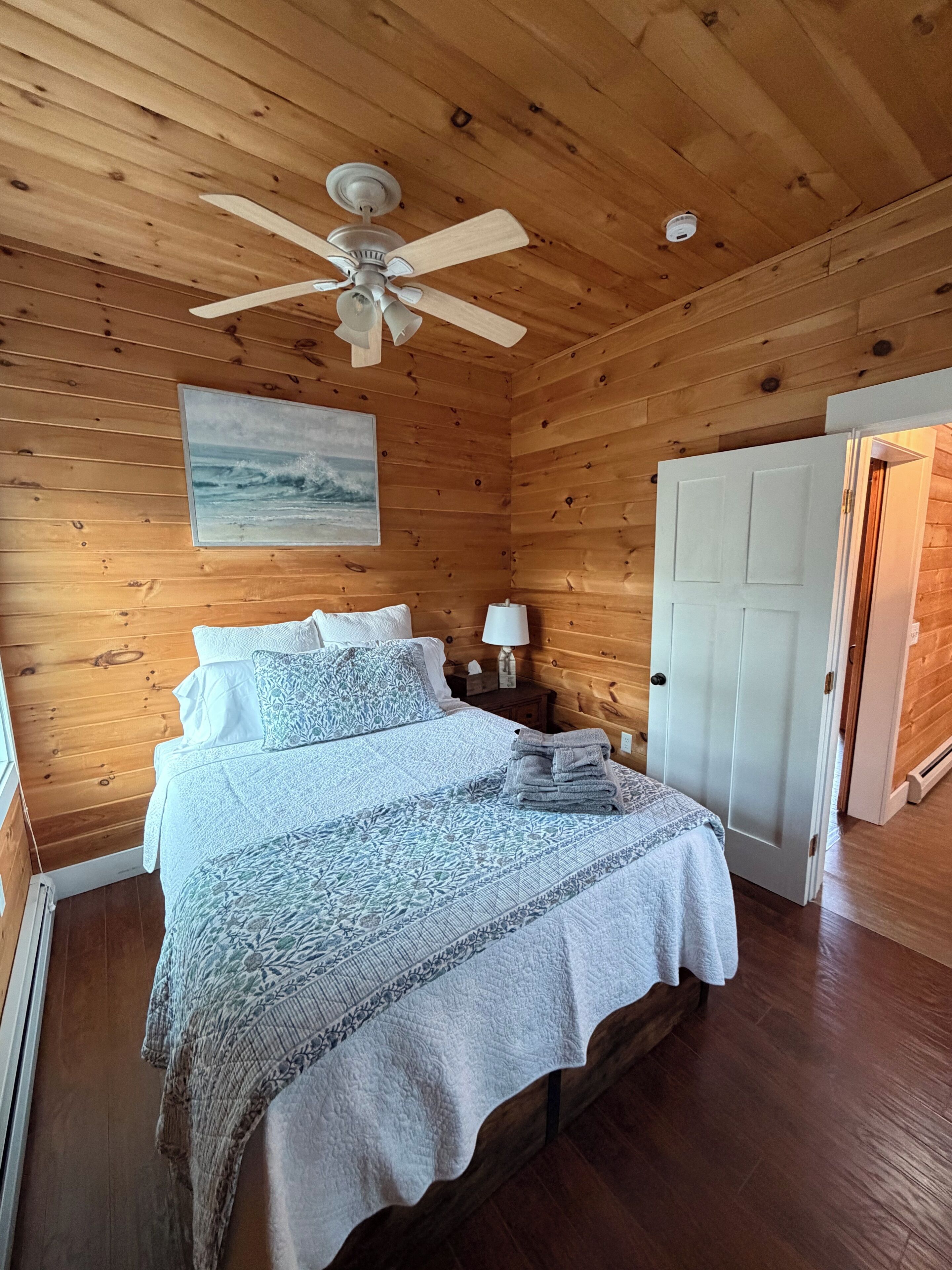 Habitación con paneles de madera, una cama, un ventilador de techo, un cuadro y una puerta.