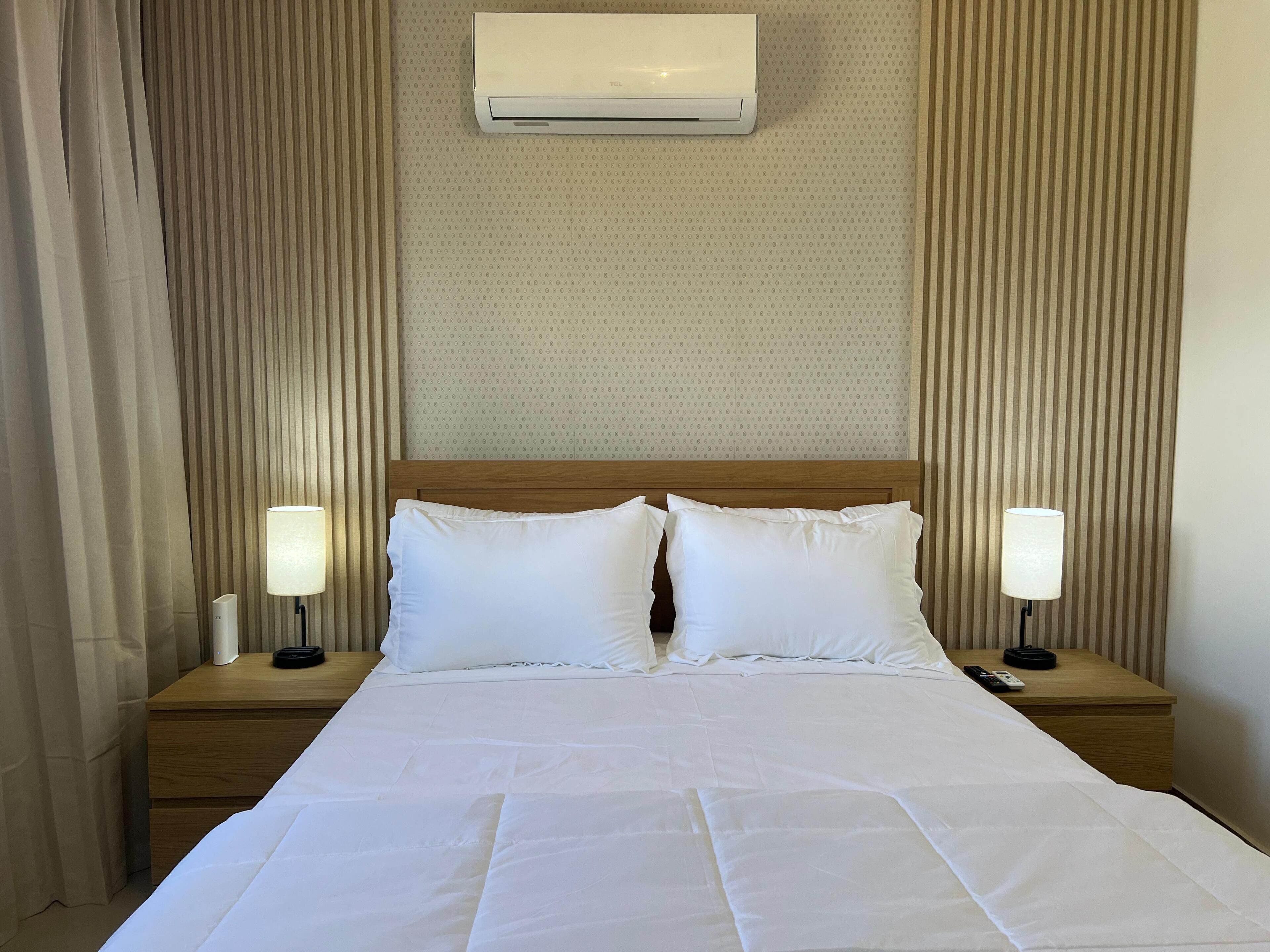 A hotel room with a bed, two bedside tables, a nightstand, an air conditioning unit, and curtains.