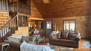 Interior de una cabaña de troncos con una escalera, un comedor, una sala de estar con sofá y sillones, y una vista a través de una ventana.