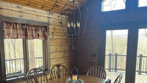 Un interior de madera con una mesa redonda para comer rodeada de sillas, una lámpara colgante y ventanas amplias con vistas a árboles.