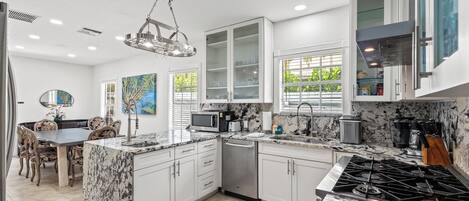A modern kitchen with white cabinetry, a central island, a dining area with a table and chairs, and a large window with blinds.