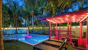 Ett poolområde med solstolar, en uppblåsbar rosa flamingo och en tiki-hydda dekorerad med ljusslingor.