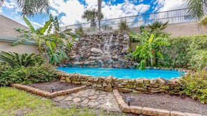 A pool with a waterfall, surrounded by stone walls and tropical plants. - Coastal Condo Oasis | Walk to Beach (Fernandina Beach)