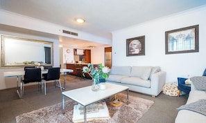 A modern living room with a sofa, coffee table, and chairs, a dining area with a table and chairs, and a kitchen in the background. - Sky-High 2BR at Chevron – Level 15 Ocean Views (Gold Coast)