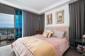 A hotel room with a large bed, a chair, a small table with books, a balcony with a view, and two framed pictures on the wall. - Towers of Chevron - 3152 (2BR/1BA) (Gold Coast)