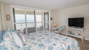 A bedroom with a bed, a TV, a chair, and a window with blinds. - Sand Dollar 1-407 Amazing Oceanfront Views from Private Balcony! (St. Augustine)