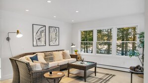 A living room with a sofa, coffee table, and two floor lamps. There are two framed artworks on the wall and a large window with a view of trees.