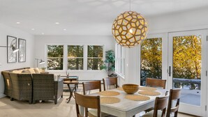 A dining area with a table, chairs, a hanging light fixture, and a view of trees through large windows.