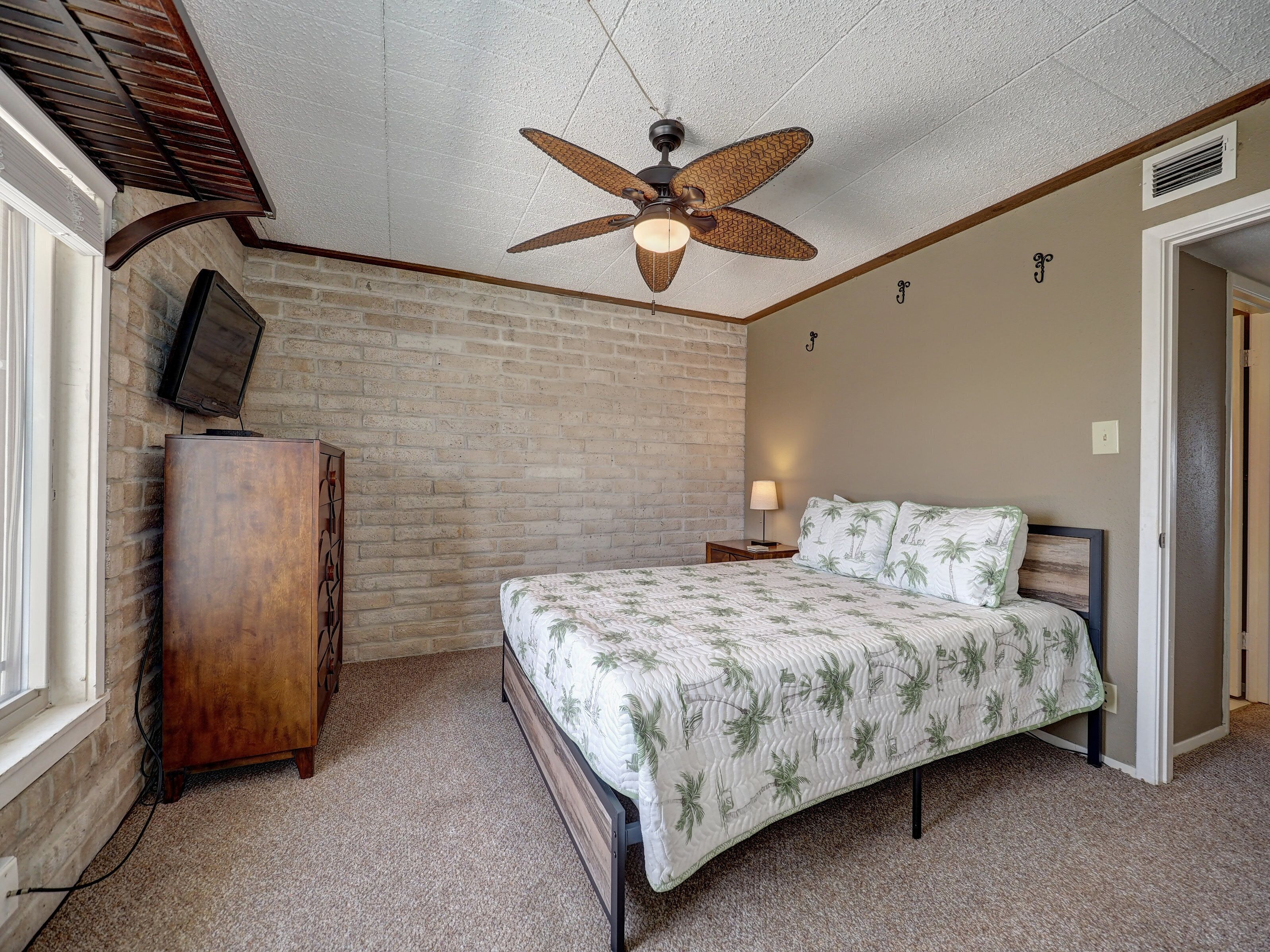 A bedroom with a bed, a wooden dresser, a ceiling fan, a TV mounted on the wall, and a door leading to another room.