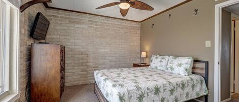 A bedroom with a bed, a wooden dresser, a ceiling fan, a TV mounted on the wall, and a door leading to another room.