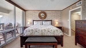 A bedroom with a large bed, a wooden bench, a dresser, and a view through a glass door to an outdoor area. - Charming Coral Cay #207: 2BR condo with pool view in Port Aransas. (Port Aransas)