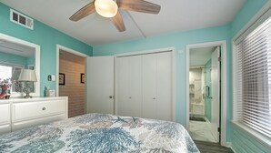 Um quarto com ventoinha no teto, cama com colcha estampada, cômoda, espelho e banheiro visível por uma porta aberta.